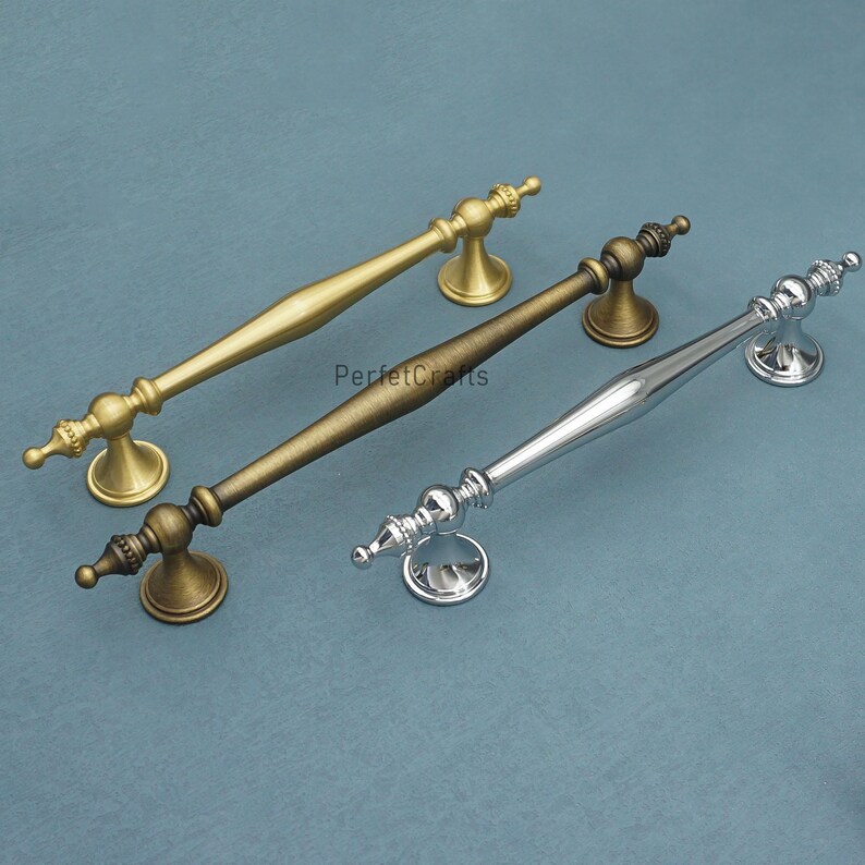 Może przedstawiać: Trzy dekoracyjne uchwyty do szafek w kolorze złotym, brązowym i srebrnym. Każdy uchwyt ma ozdobny design z zakrzywionym prętem i dekoracyjnymi końc&oacute;wkami. Uchwyty są prezentowane na niebiesko-zielonej powierzchni. Widoczny jest tekst "PerfectCrafts".