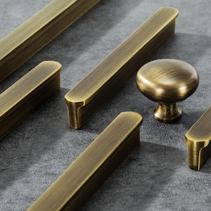 Puede incluir: Primer plano de herrajes de gabinete de latón antiguo, que incluyen tiradores de barra largos y un pomo redondo. Los herrajes tienen un acabado cálido y envejecido y están dispuestos sobre una superficie gris. El texto "Perfect Craft" es visible en el pomo.