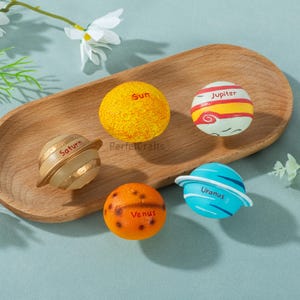 May include: A collection of colorful, spherical planet models, including the Sun, Saturn, Jupiter, Venus, and Uranus. Each planet is painted with detailed features and labeled with its name. The planets are arranged on a wooden tray.