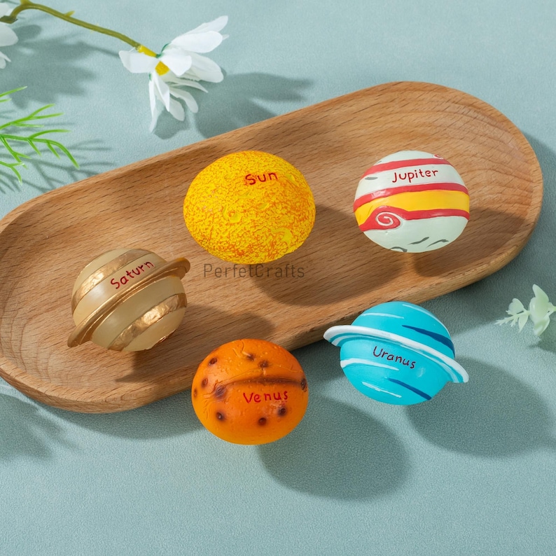 May include: A collection of colorful, spherical planet models, including the Sun, Saturn, Jupiter, Venus, and Uranus. Each planet is painted with detailed features and labeled with its name. The planets are arranged on a wooden tray.