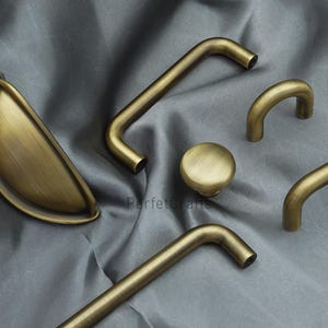 K&ouml;nnte beinhalten: Eine Auswahl an antiken Messing-Schrankbeschl&auml;gen, darunter ein Muschelgriff, ein runder Knopf und mehrere Stangen- und gebogene Griffe. Die Beschl&auml;ge sind auf einem grauen Stoff arrangiert, der die verschiedenen Formen und Designs zeigt.