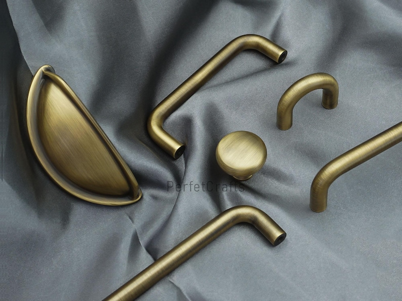 K&ouml;nnte beinhalten: Eine Auswahl an antiken Messing-Schrankbeschl&auml;gen, darunter ein Muschelgriff, ein runder Knopf und mehrere Stangen- und gebogene Griffe. Die Beschl&auml;ge sind auf einem grauen Stoff arrangiert, der die verschiedenen Formen und Designs zeigt.