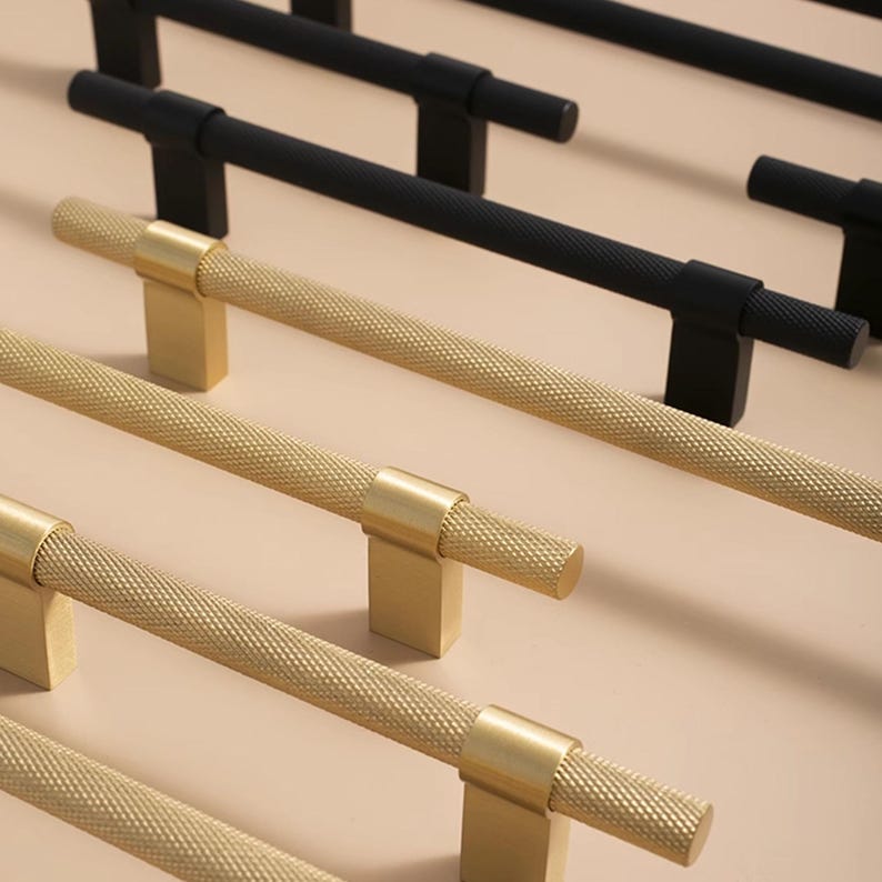 K&ouml;nnte beinhalten: Nahaufnahme von Schrankbeschl&auml;gen. Das Bild zeigt mehrere goldene und schwarze Schrankgriffe. Die goldenen Griffe haben ein strukturiertes Rautenmuster auf der Stange. Die schwarzen Griffe haben eine glatte, matte Oberfl&auml;che. Die Griffe sind in parallelen Reihen angeordnet.