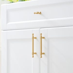 May include: Close-up of a white cabinet with gold-coloured hardware. The cabinet has a drawer with a small knob and two doors with long, vertical handles. The hardware has a classic, elegant design.