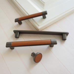 May include: A collection of cabinet handles and a knob. The handles have a dark grey metal base with brown leather wrapping. The knob and handles are displayed on a white surface. The text "PerfecCrafts" is visible on the image.
