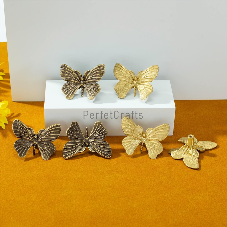 May include: A collection of bronze and gold-colored butterfly-shaped cabinet pulls. The pulls are in various stages of being assembled. The image is set against a white background and a mustard-colored surface.