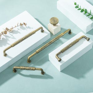 Può includere: Un assortimento di ferramenta per mobili in ottone antico, tra cui maniglie e un pomello. Le maniglie variano in dimensioni e presentano un design martellato e strutturato. L'hardware è esposto su piattaforme bianche su uno sfondo azzurro. Misure in centimetri.