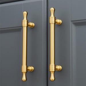 May include: Two gold-coloured cabinet pulls with a vertical bar design. The pulls are mounted on a grey cabinet door. The pulls have a cylindrical body with decorative finials at the top and bottom.