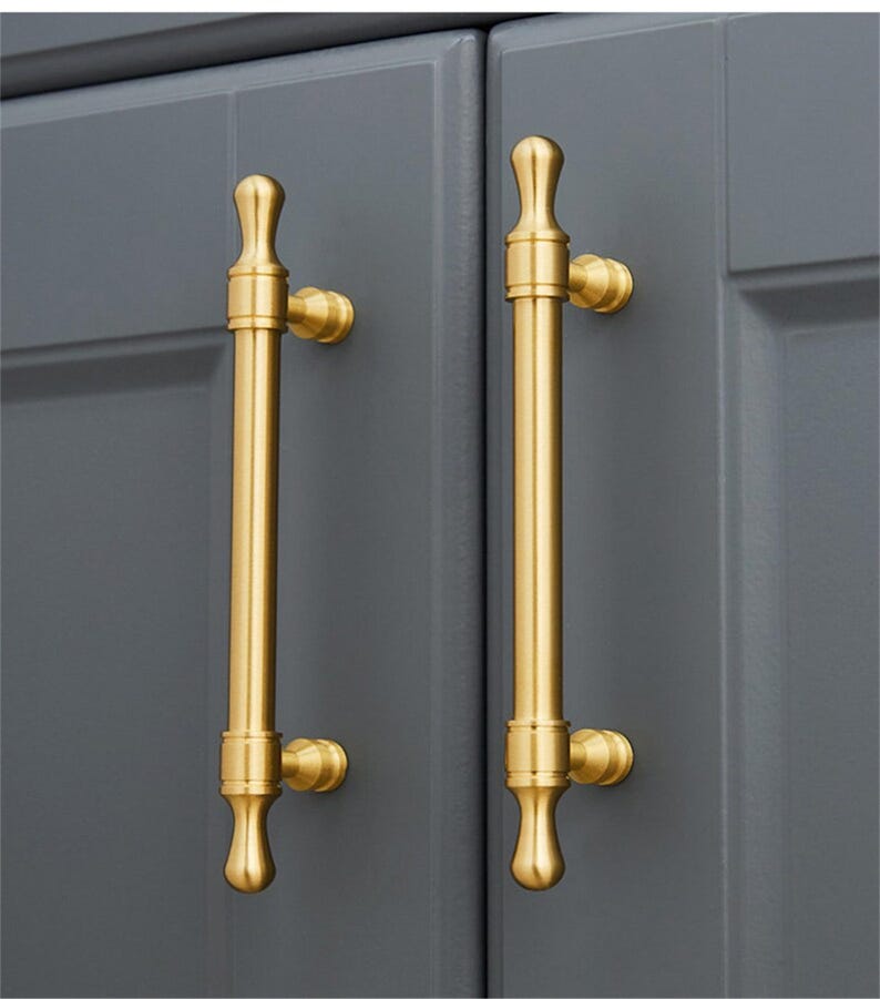 May include: Two gold-coloured cabinet pulls with a vertical bar design. The pulls are mounted on a grey cabinet door. The pulls have a cylindrical body with decorative finials at the top and bottom.