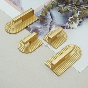 May include: Four gold-colored cabinet pulls with a retro design. Each pull features a rectangular handle and a base with a series of curved and straight lines. The pulls are arranged on a light-colored surface.