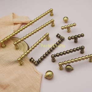 Op de afbeelding: Een verzameling antieke messing kastbeslagen, waaronder handgrepen en knoppen. De handgrepen hebben een kralenontwerp en de knoppen zijn bolvormig. De hardware is verkrijgbaar in verschillende maten en tinten messing en brons.