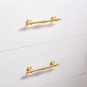 Może przedstawiać: Dwa złote uchwyty do szafek zamontowane na białej powierzchni. Każdy uchwyt ma zakrzywioną konstrukcję pręta z dekoracyjnymi końcami. Uchwyty są ustawione poziomo, co sugeruje, że są to uchwyty do szuflad lub drzwi.