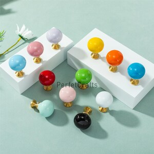 May include: A selection of brightly coloured spherical cabinet knobs. The knobs are in shades of blue, pink, red, green, yellow, and white. Each knob has a gold-coloured base. The text "PerfetcCrafts" is visible in the image.
