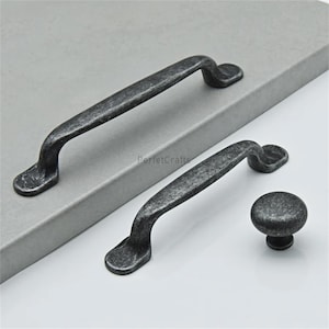 May include: Two dark grey cabinet pulls and a matching knob. The pulls have a curved handle design with rounded ends. The knob is a small, round shape. The hardware has a distressed, antique finish.