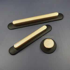 May include: A set of black and gold cabinet hardware. The set includes two long, rectangular pulls and a round knob. The pulls have a black base with a gold, textured centre. The knob has a matching gold centre. The hardware is displayed on a grey surface.