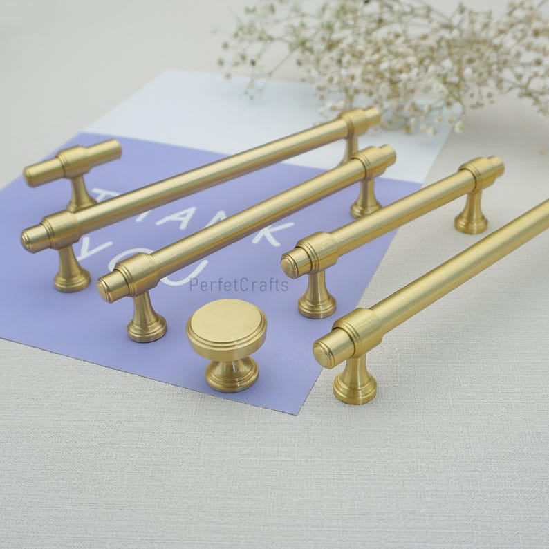 Peut inclure: Ensemble de quincaillerie de meubles dor&eacute;e, comprenant plusieurs poign&eacute;es de tirage et un bouton rond. Le mat&eacute;riel est dispos&eacute; sur une surface violet clair avec le texte "PerfektCrafts" visible. Le mat&eacute;riel a un design classique et &eacute;l&eacute;gant.