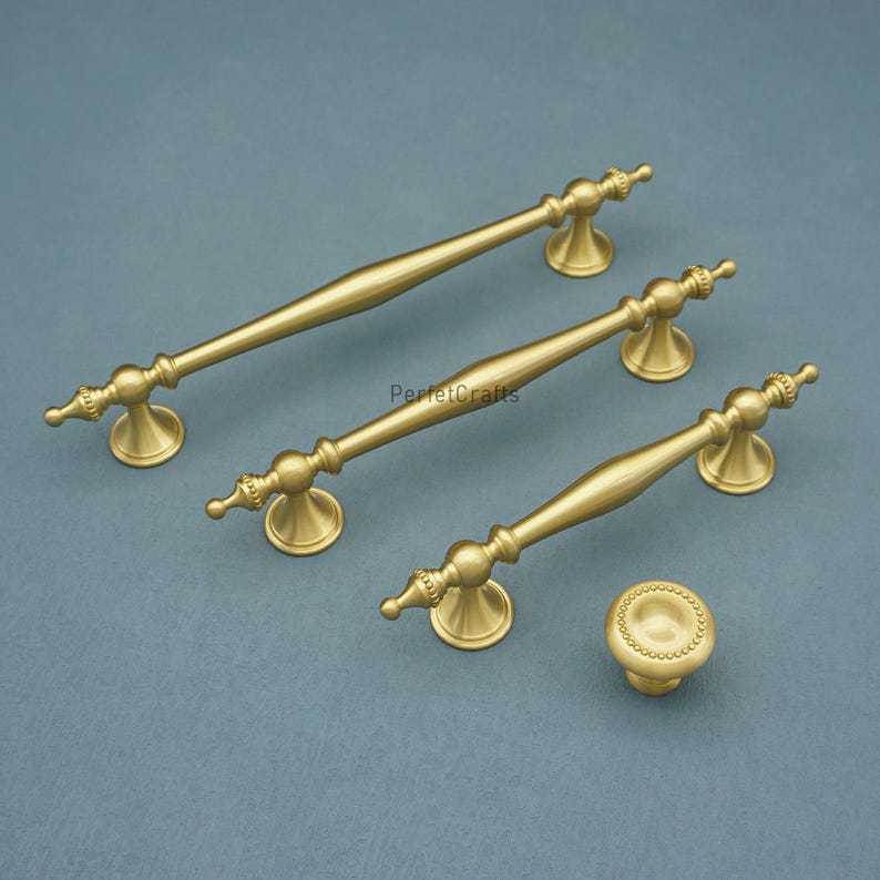 Może przedstawiać: Zestaw czterech złotych element&oacute;w okuć do szafek. Zestaw zawiera trzy podłużne uchwyty i jeden okrągły gałkę. Uchwyty mają dekoracyjny design z zakrzywionym kształtem i są zamontowane na niebiesko-szarej powierzchni. Widoczny jest tekst "PerfecCrafts".