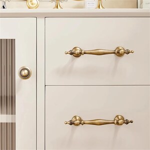 Może przedstawiać: Zbliżenie kremowej szafki z antycznym mosiężnym osprzętem. Szafka posiada dwie szuflady z pasującymi uchwytami i okrągłą gałką na drzwiach. Uchwyty są ozdobne, z dekoracyjnymi detalami.