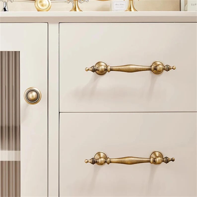 Może przedstawiać: Zbliżenie kremowej szafki z antycznym mosiężnym osprzętem. Szafka posiada dwie szuflady z pasującymi uchwytami i okrągłą gałką na drzwiach. Uchwyty są ozdobne, z dekoracyjnymi detalami.