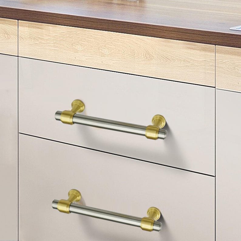 Peut inclure: Deux poign&eacute;es de tiroir d'armoire avec une barre argent&eacute;e bross&eacute;e et des embouts dor&eacute;s. Les poign&eacute;es sont mont&eacute;es sur une porte d'armoire de couleur claire. Le meuble a un comptoir en bois.