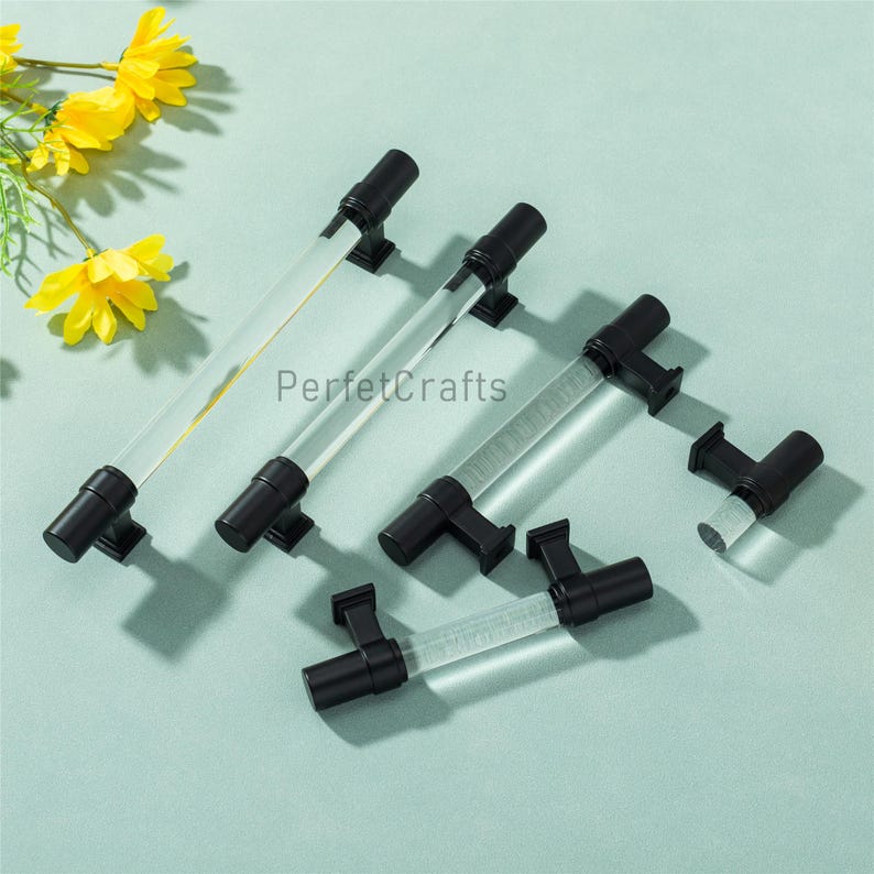 May include: A collection of clear acrylic cabinet pulls with black metal hardware. The pulls have a cylindrical design and are arranged on a light blue surface. The text "PerfetcCrafts" is visible on the pulls.