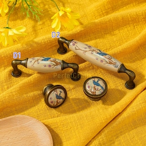 May include: A set of decorative cabinet hardware featuring a bird and floral design. The set includes two handles and two knobs. The handles are cream-colored with a bronze-colored base. The knobs are round with a similar design.