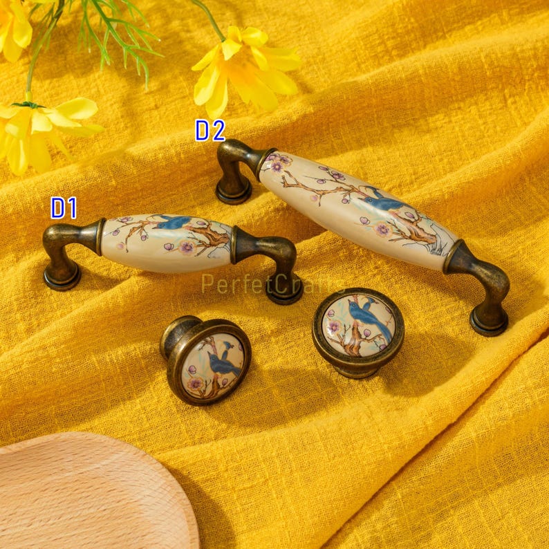 May include: A set of decorative cabinet hardware featuring a bird and floral design. The set includes two handles and two knobs. The handles are cream-colored with a bronze-colored base. The knobs are round with a similar design.