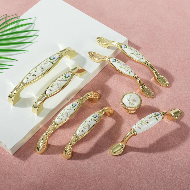 Pu&ograve; includere: Un assortimento di maniglie dorate per mobili e un pomello. Presentano un intarsio in ceramica bianca con un delicato motivo floreale blu, verde e viola. Le maniglie hanno un design curvo.