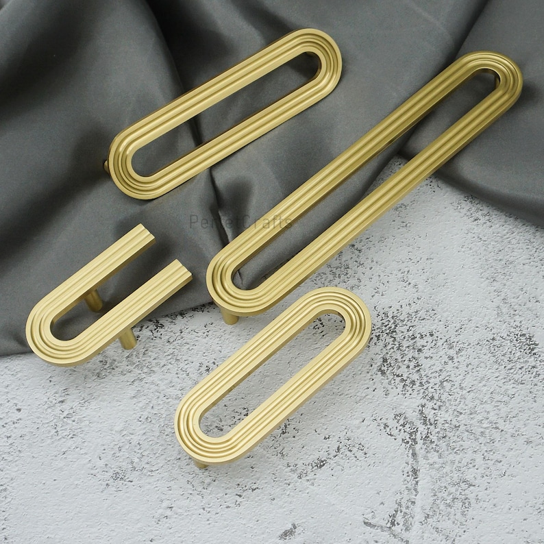 May include: Four gold-coloured cabinet pulls with a grooved, oval design. The pulls are arranged on a grey and white surface. The hardware has a modern, minimalist aesthetic, suitable for drawers or cabinets.
