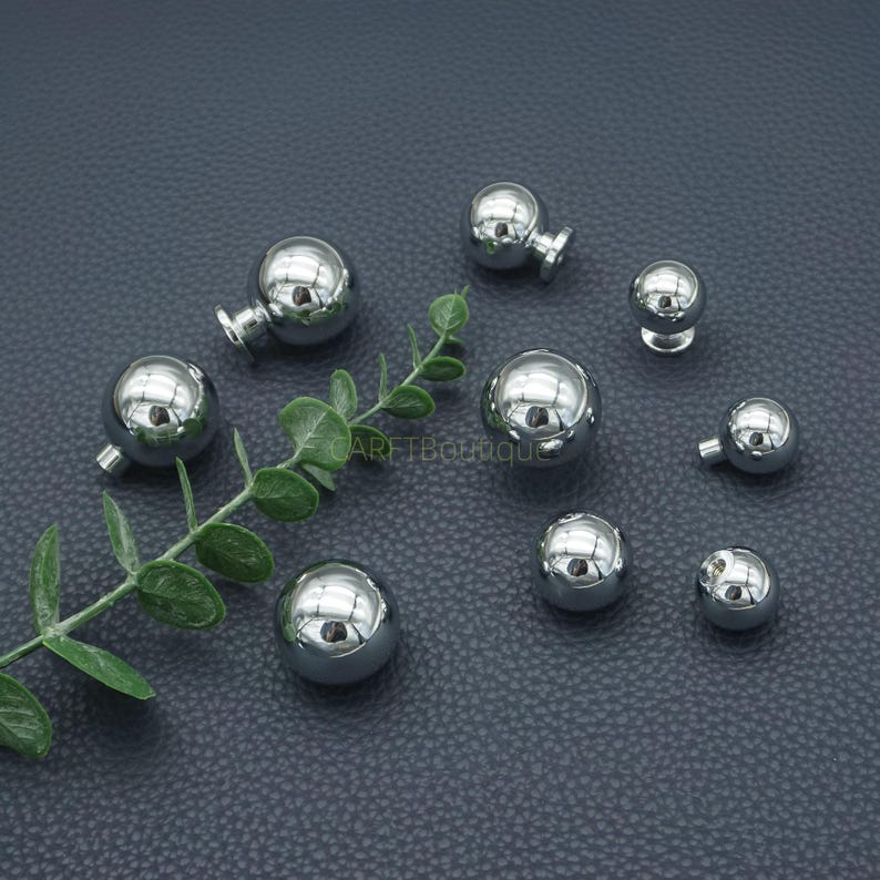 Puede incluir: Una colecci&oacute;n de pendientes de bola de metal plateado brillante de varios tama&ntilde;os. Algunos tienen un peque&ntilde;o poste. Las superficies reflectantes crean un efecto de espejo. Una ramita de hojas verdes est&aacute; en primer plano.