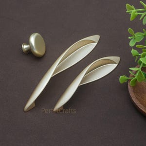 以下が含まれることがあります： ゴールドトーンのキャビネットハードウェアのセット。2つの曲線ハンドルと丸いノブが含まれています。ハンドルは、葉のような流れるようなデザインです。ハードウェアは茶色の表面に表示されています。