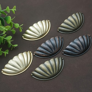 Könnte beinhalten: Sechs dekorative Schubladengriffe in verschiedenen metallischen Ausführungen, darunter Gold, Bronze und Dunkelgrau. Jeder Griff hat ein fächerförmiges Design mit geriffelter Textur. Die Griffe sind auf einer dunkelbraunen Oberfläche angeordnet, mit einer grünen Pflanze im Hintergrund.