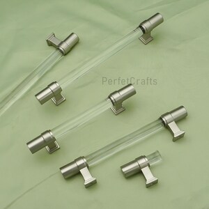 May include: Clear acrylic cabinet pulls with brushed nickel hardware. The pulls have a cylindrical design and are mounted on square bases. The text "PerfecCrafts" is visible in the image.
