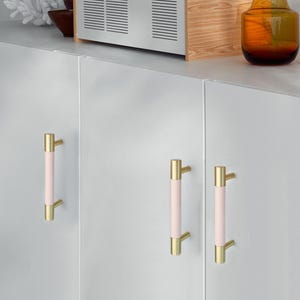 Puede incluir: Primer plano de tres tiradores de armario. Cada tirador tiene una secci&oacute;n central rosa claro y extremos dorados. Los tiradores est&aacute;n montados en una puerta de armario blanca. Los tiradores miden aproximadamente 13 cm de largo.