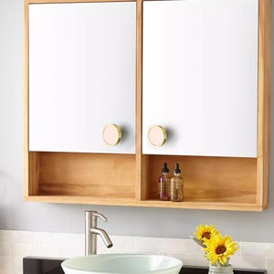 Puede incluir: Un mueble de ba&ntilde;o de madera con dos puertas con espejo y una balda inferior. Las puertas tienen pomos redondos dorados con un centro rosa. El mueble est&aacute; montado sobre un lavabo con un grifo cromado. Dos botellas est&aacute;n en el estante.