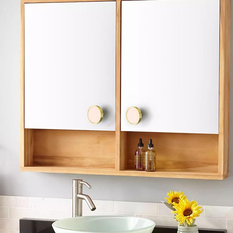 Puede incluir: Un mueble de ba&ntilde;o de madera con dos puertas con espejo y una balda inferior. Las puertas tienen pomos redondos dorados con un centro rosa. El mueble est&aacute; montado sobre un lavabo con un grifo cromado. Dos botellas est&aacute;n en el estante.