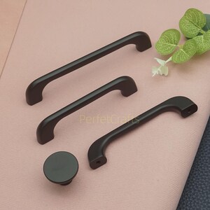 Op de afbeelding: Een verzameling donkerbruine kastbeslagen, waaronder drie handgrepen van verschillende afmetingen en een ronde knop. De handgrepen hebben een gebogen ontwerp en zijn gemaakt van een materiaal met een matte afwerking. De tekst "PerfecCrafts" is zichtbaar.