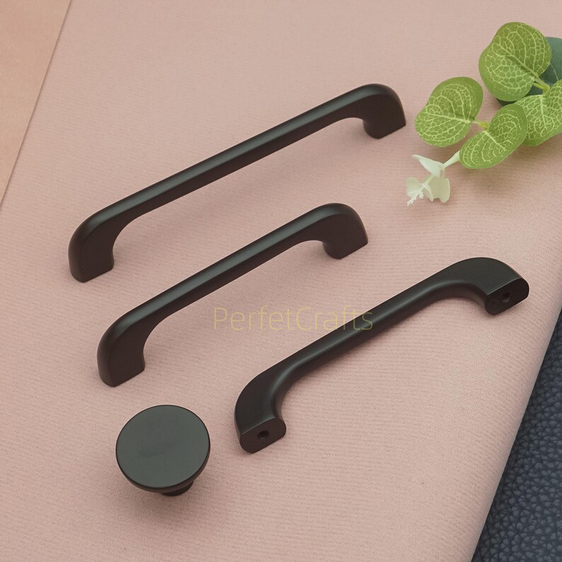 Op de afbeelding: Een verzameling donkerbruine kastbeslagen, waaronder drie handgrepen van verschillende afmetingen en een ronde knop. De handgrepen hebben een gebogen ontwerp en zijn gemaakt van een materiaal met een matte afwerking. De tekst "PerfecCrafts" is zichtbaar.