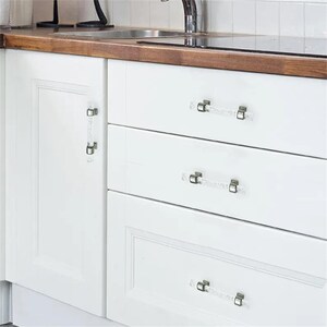 Peut inclure: Des armoires de cuisine blanches avec des poign&eacute;es de tiroir transparentes en forme de cristal. Les poign&eacute;es sont rectangulaires avec des accents m&eacute;talliques argent&eacute;s. Les armoires font partie d'une cuisine avec un plan de travail en bois et un &eacute;vier en acier inoxydable.