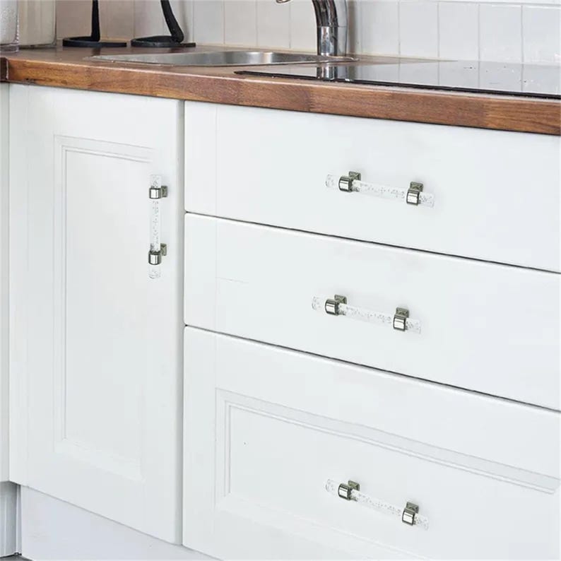 Peut inclure: Des armoires de cuisine blanches avec des poign&eacute;es de tiroir transparentes en forme de cristal. Les poign&eacute;es sont rectangulaires avec des accents m&eacute;talliques argent&eacute;s. Les armoires font partie d'une cuisine avec un plan de travail en bois et un &eacute;vier en acier inoxydable.