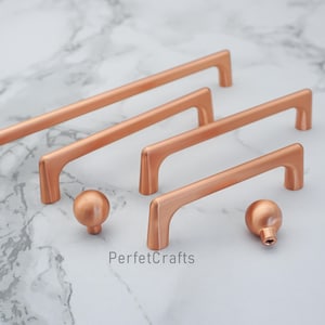 May include: Copper-colored cabinet hardware set, including several bar pulls and two round knobs. The pulls have a curved design and are of varying lengths. The knobs and pulls are displayed on a marble-patterned surface. The text "PerfecCrafts" is visible.