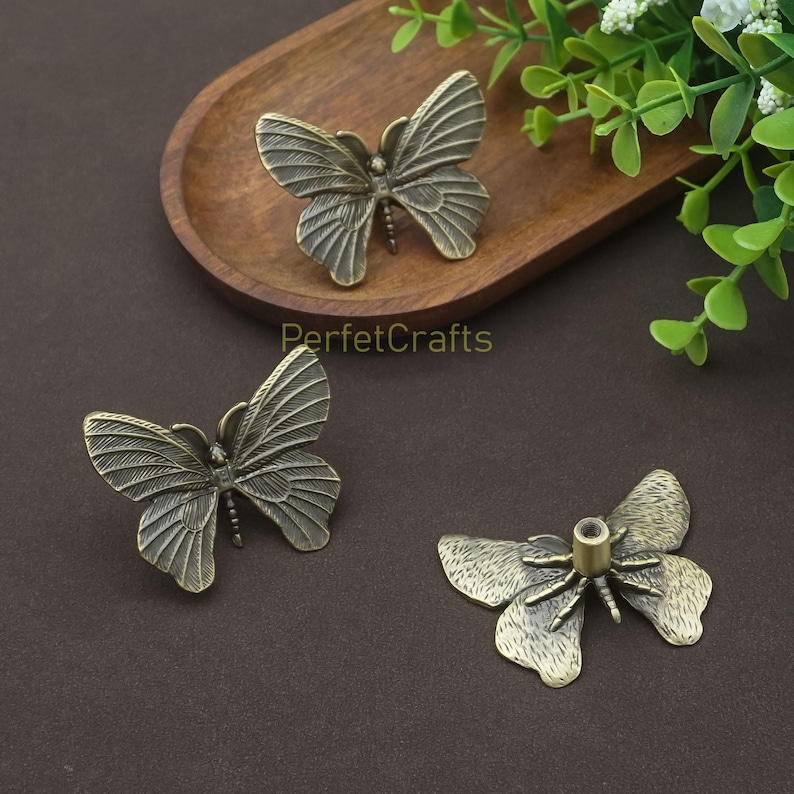 May include: Three antique bronze butterfly-shaped cabinet knobs. The knobs have detailed wing patterns and are displayed on a wooden tray and a brown surface. The text "PerfctCrafts" is visible.