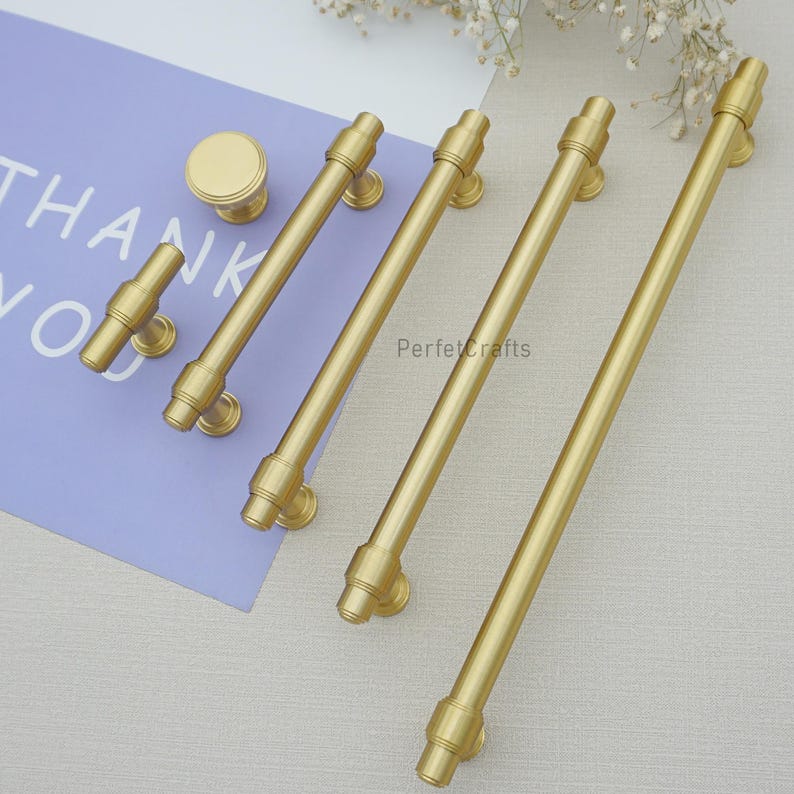 Peut inclure: Ensemble de quincaillerie de placard dor&eacute;e, comprenant des boutons et des barres de tirage. Le mat&eacute;riel a un design cylindrique avec des d&eacute;tails d&eacute;coratifs. Le mot "PerfecCrafts" est visible. L'ensemble est dispos&eacute; sur une surface claire.