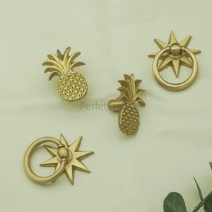 May include: Four gold-coloured cabinet hardware pieces. Two are pineapple-shaped knobs, and two are ring pulls with starburst backplates. The hardware is displayed on a light background. The text "Perfect Craft" is visible on the image.