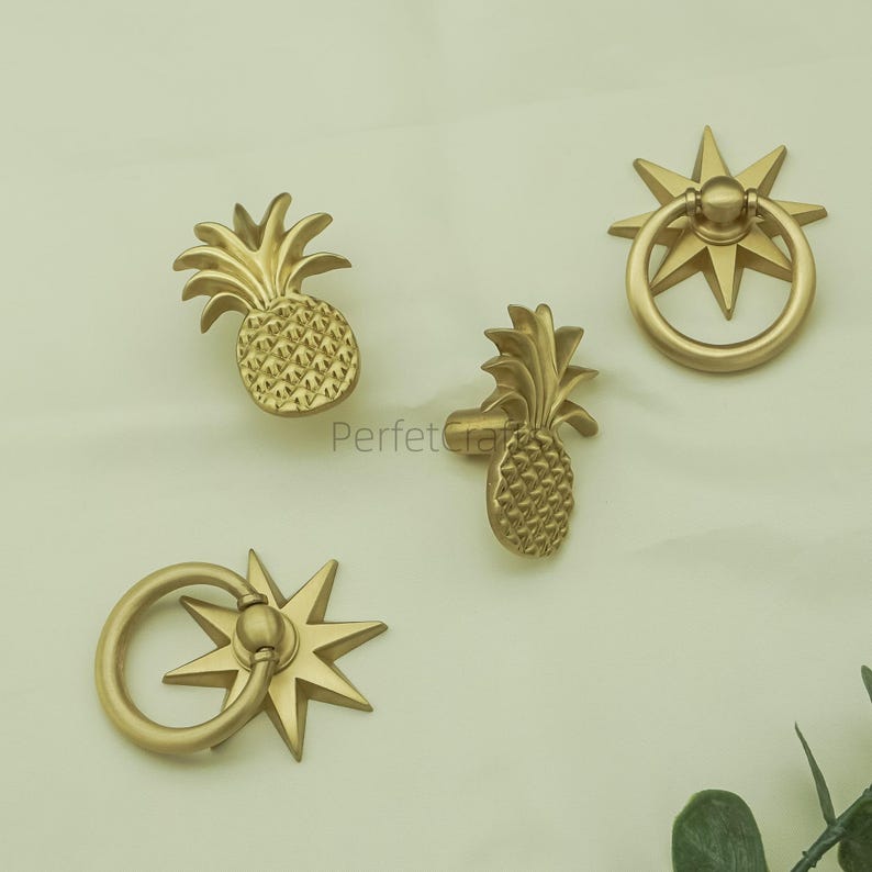 May include: Four gold-coloured cabinet hardware pieces. Two are pineapple-shaped knobs, and two are ring pulls with starburst backplates. The hardware is displayed on a light background. The text "Perfect Craft" is visible on the image.