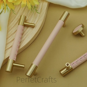 Puede incluir: Un conjunto de herrajes para armarios, que incluye tiradores y un pomo. Los herrajes presentan un centro rosa claro con detalles dorados. Un tirador tiene un dise&ntilde;o cosido. El texto "PerfetcCrafts" es visible en la parte inferior.