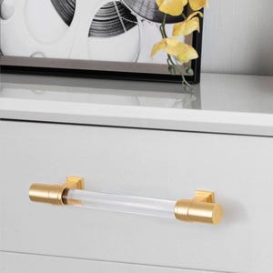 May include: A clear acrylic and gold-toned metal drawer pull. The cylindrical acrylic center is flanked by gold-colored metal end caps and mounting brackets. The hardware is mounted on a white drawer front.
