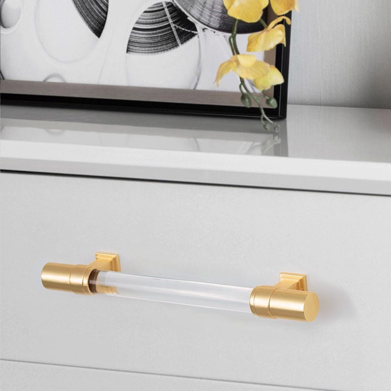 May include: A clear acrylic and gold-toned metal drawer pull. The cylindrical acrylic center is flanked by gold-colored metal end caps and mounting brackets. The hardware is mounted on a white drawer front.