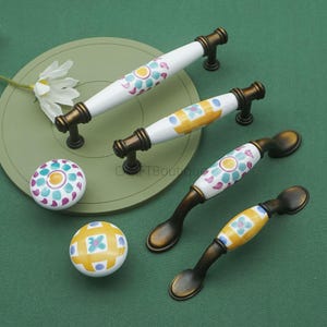 May include: A collection of decorative cabinet hardware, including white ceramic handles and knobs with colorful floral patterns. The handles have a bronze finish and are arranged on a green surface.