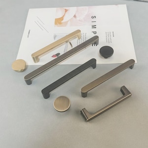 May include: A selection of cabinet handles and knobs in gold, bronze, and black finishes. The image shows a variety of bar pulls and round knobs. The word "SIMPLE" is visible on a magazine page. The hardware is displayed on a neutral background.